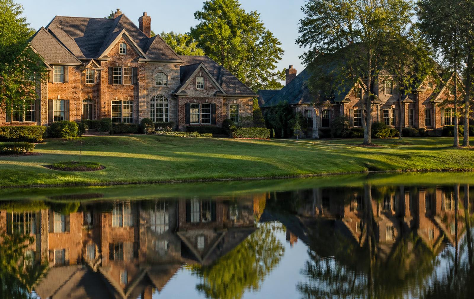 Large brick homes with deep lawns backing onto a still lake at golden hour in a South Charlotte golf community