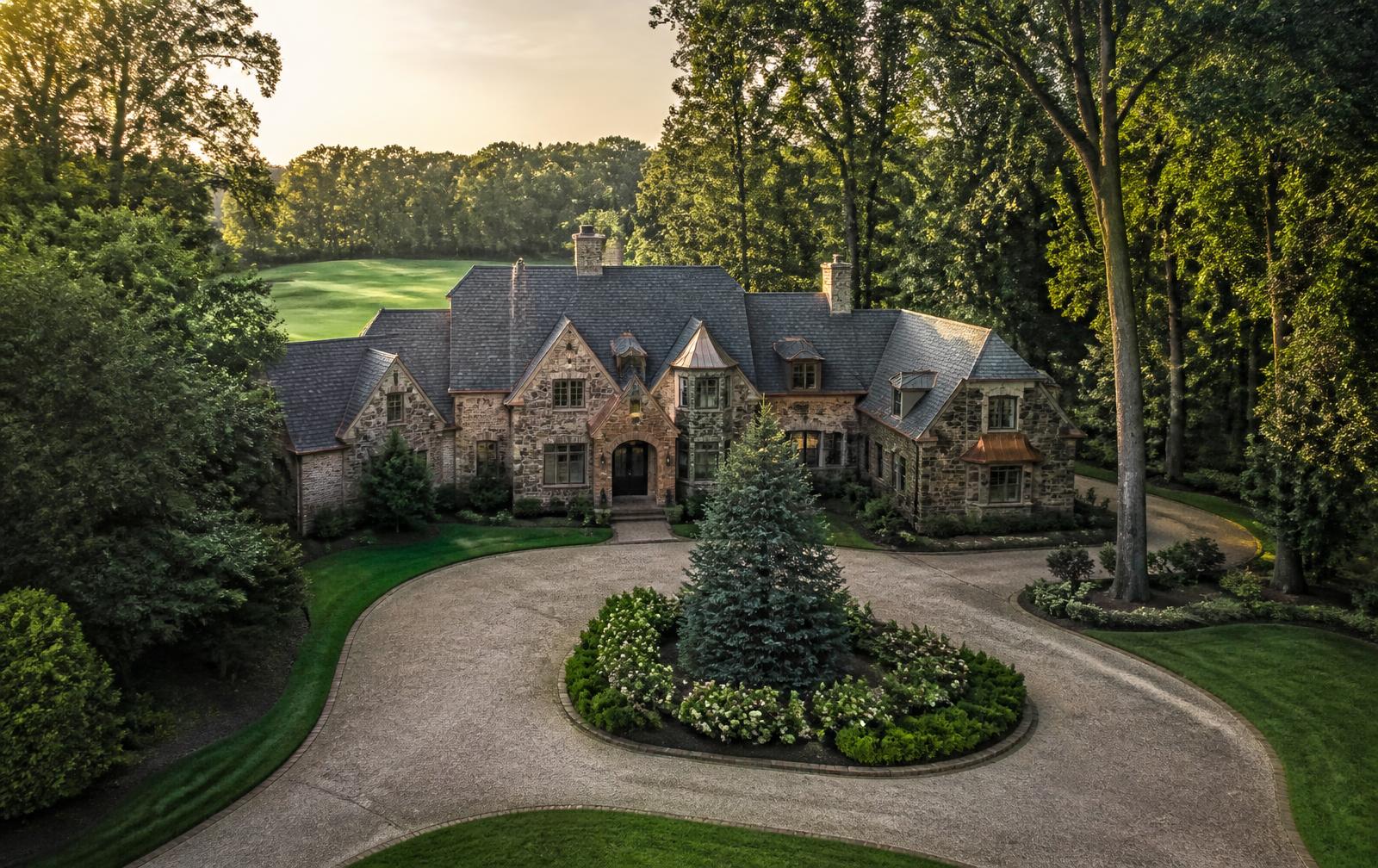 Large custom stone and brick estate home set back on a deeply wooded lot with a manicured fairway visible beyond the tree line